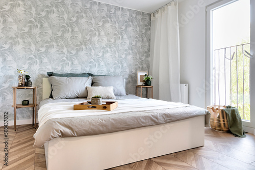 Modern interior of bedroom with window, wooden floor and comfortable double bed with pillows sheets and blanket. Cozy interior for rest and trendy design.