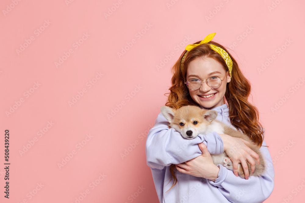 Cheerful redhead young girl with long curly hair holding cute little puppy of corgi dog isolated on pink background. Concept of youth, beauty, lifestyle Cheerful redhead young girl with long curly hair holding cute little puppy of corgi dog isolated on pink background. Concept of youth, beauty, lifestyle