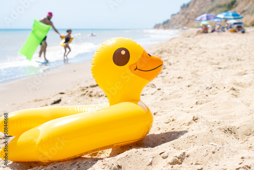 Yellow inflatable duck ring laying on sandy beach near blue wavy ocean in sunny day with people and sun umbrellas on background. Protection swim tube for kid. Summer travel vacation resort concept