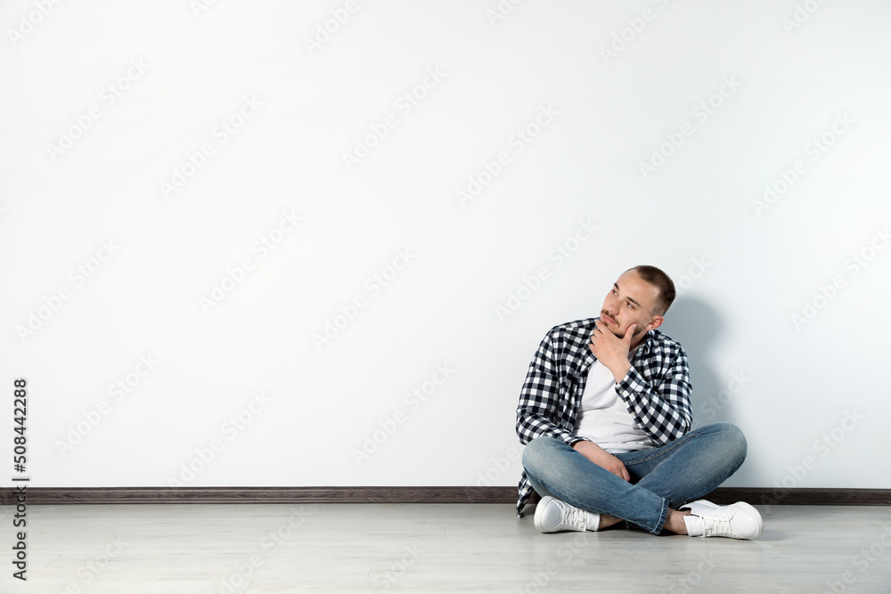 Handsome young man sitting on floor near white wall indoors, space for text Handsome young man sitting on floor near white wall indoors, space for text