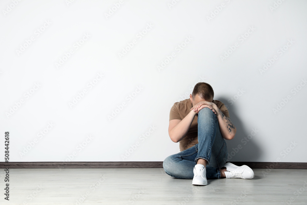 Sad young man sitting on floor near white wall indoors, space for text Sad young man sitting on floor near white wall indoors, space for text