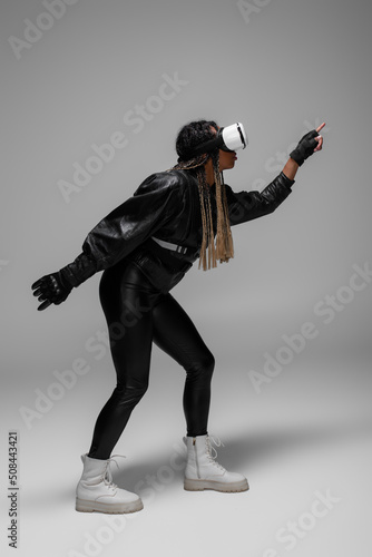 Side view of african american woman in virtual reality headset and leather jacket gaming on grey background.