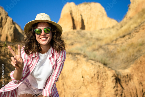 Happy young woman tourist enjoying vacation adventure, standing on yellow sandy mountains and blue sky background in grand canyon, in sunglasses and summer hat smiling looking at camera. Copy space