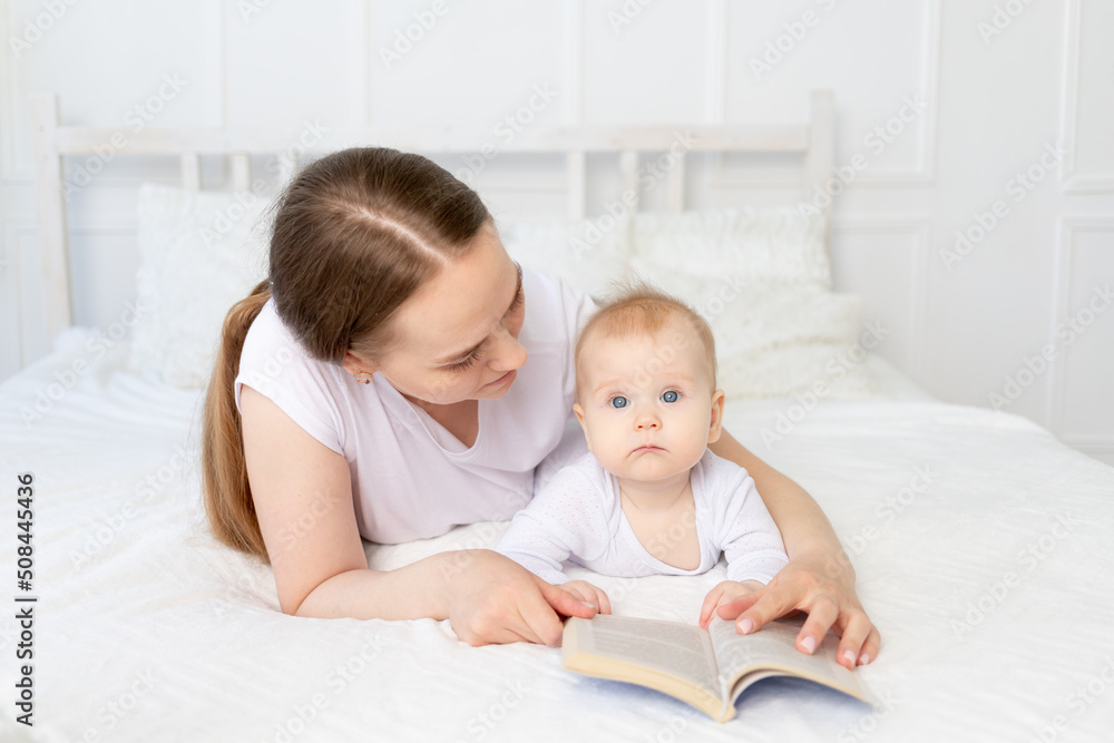 mom reads a book to baby before going to bed on a white bed with cotton bedding at home, happy family mom reads a book to baby before going to bed on a white bed with cotton bedding at home, happy family