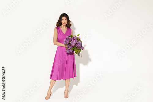 portrait of elegant woman wearing purple dress and holding lilac brunches against white wall. International woman’s day, Mother’s day and spring concept.