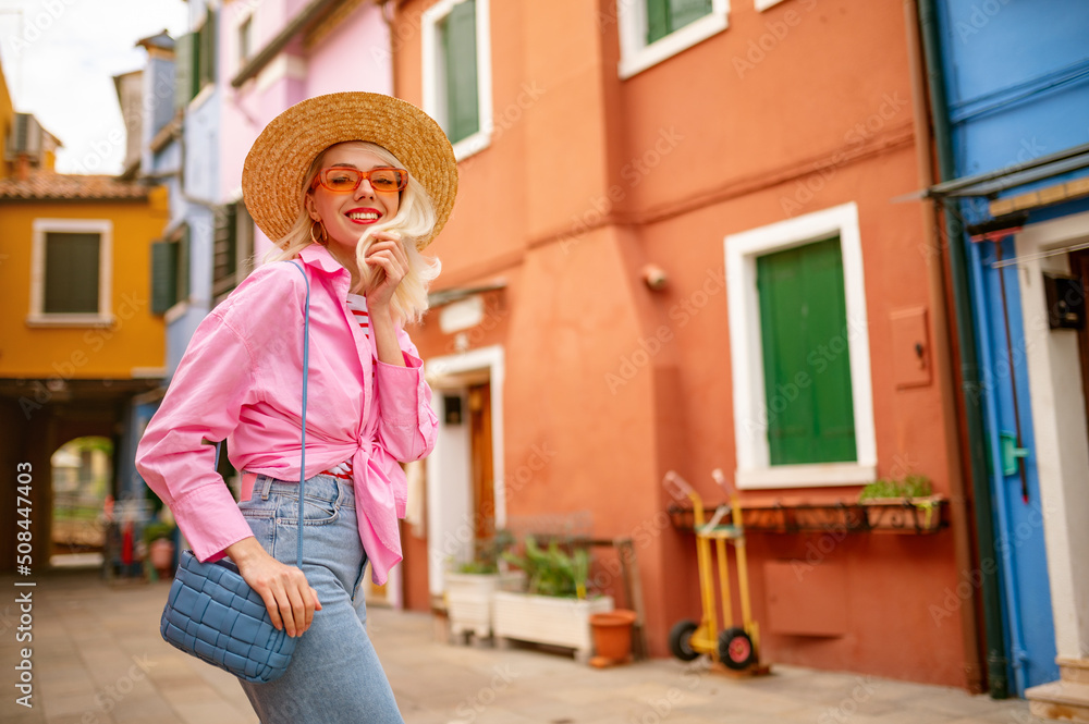 Happy smiling traveler woman posing in street, among colorful houses. Model wearing trendy summer outfit with orange sunglasses, straw hat, pink shirt, shoulder blue bag. Copy, empty space for text