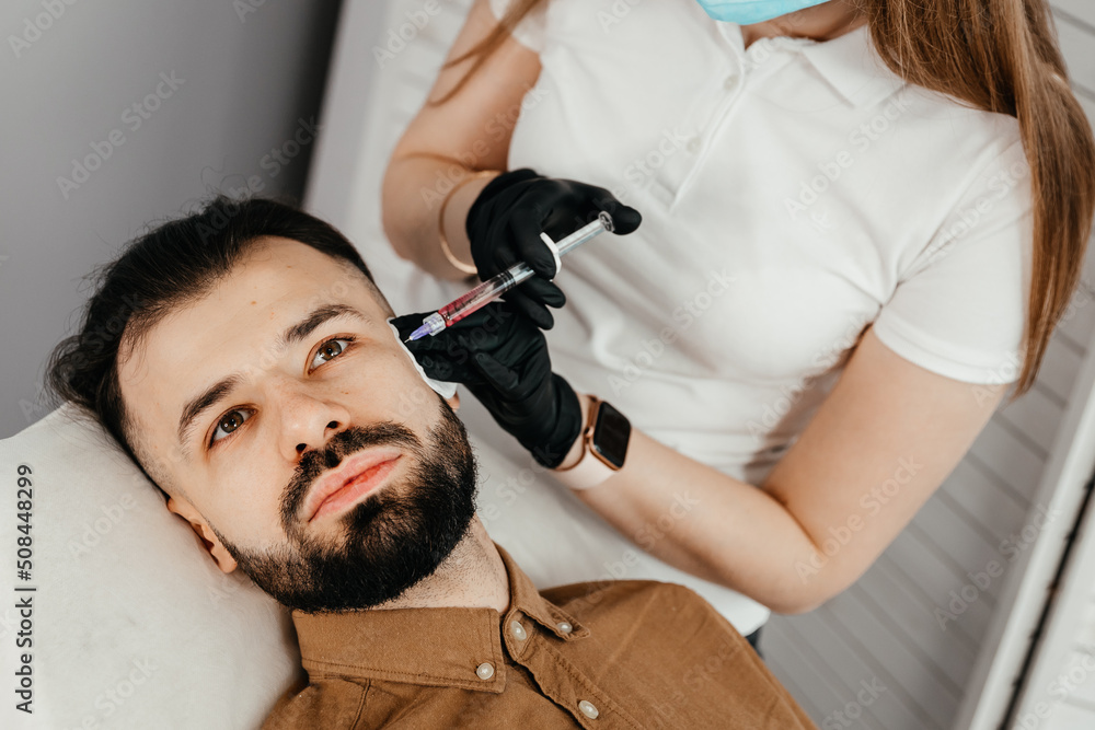 Fototapeta premium Stylish bearded man visiting aesthetic clinic, getting lips filler, closeup. Man having beauty injection at male spa salon. Anti-aging treatment for men concept