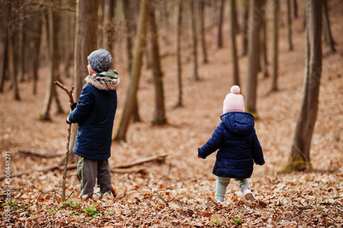 Young boy wear jacket and hat with sister in early spring forest.