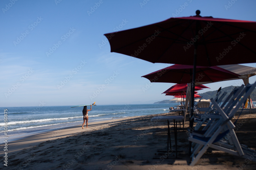 Mujer caminando con tabla de surf en la arena, sombrillas rojas y el cielo azul, playa de Sayulita, Jalisco, México, mujer surfista en traje de baño, mar y sol, deporte acuático Mujer caminando con tabla de surf en la arena, sombrillas rojas y el cielo azul, playa de Sayulita, Jalisco, México, mujer surfista en traje de baño, mar y sol, deporte acuático