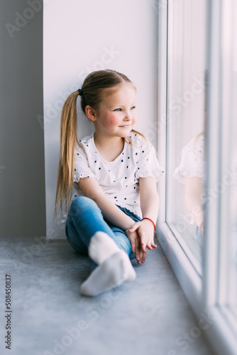Beautiful little girl smiling and watching out the window. A child looks out the window. Young girl looking from window at windowsill.
