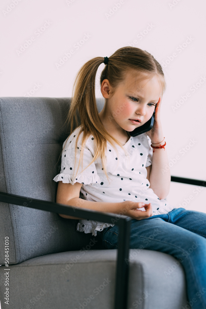 Young little confused girl talk phone sit on chair isolated on white background Young little confused girl talk phone sit on chair isolated on white background