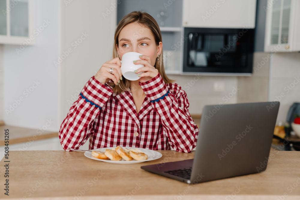 Young white happy woman having breakfast and using laptop in kitchen Young white happy woman having breakfast and using laptop in kitchen