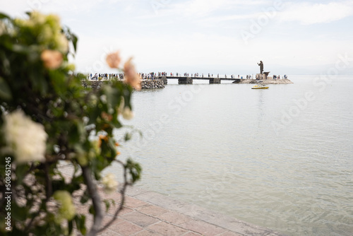 gente caminando en el Malecón del lago de Chapala, Jalisco, México, con la estatua de Jesús Cristo el pescador, un arbusto con flores y cielo nublado.