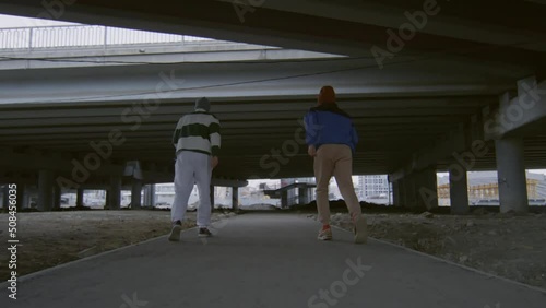 Following slow motion shot of two professional parkourists running under urban bridge and performing side flips together photo