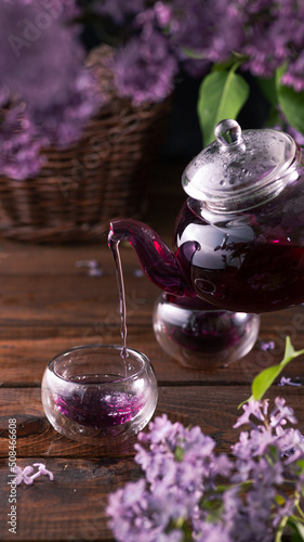 Purple tea is poured from a glass teapot into cups. Lilac flowers are all around