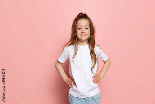 Studio shot of cute little girl, kid wearing white t-shirt posing isolated on pink background. Concept of children emotions, fashion, beauty, school and ad concept