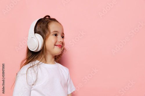 Beautiful cute little girl, kid wearing white t-shirt and headphones posing isolated on pink background. Concept of children emotions, fashion, beauty, school and ad concept