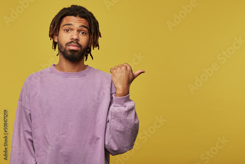 Studio portrait of young african american man posing over studio background points with a finger aside at copy space with emotionless facial expression photo