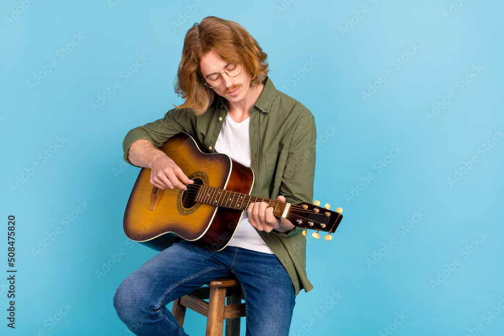 Portrait of attractive talented guy sitting on stool playing string guitar isolated over pastel blue color background Portrait of attractive talented guy sitting on stool playing string guitar isolated over pastel blue color background