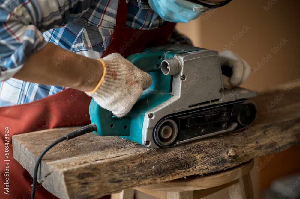 A young Asian carpenter with a brown apron uses a wood sander and tape measure while working on a plank. Carpenter concept working in a wood factory. Selective focus. A young Asian carpenter with a brown apron uses a wood sander and tape measure while working on a plank. Carpenter concept working in a wood factory. Selective focus.