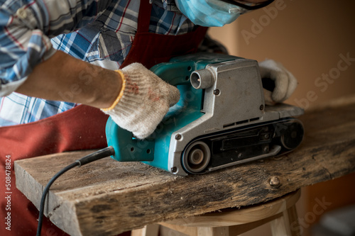 A young Asian carpenter with a brown apron uses a wood sander and tape measure while working on a plank. Carpenter concept working in a wood factory. Selective focus.