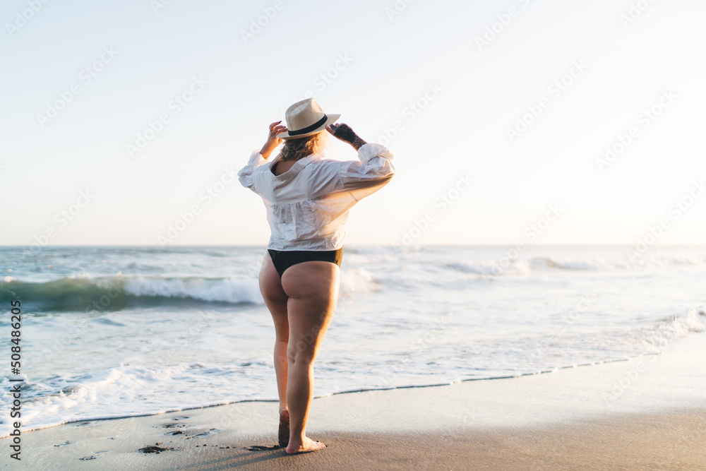 Caucasian woman in black swimsuit and white shirt standing at sandy beach of Mauritius island exploring tropical nature environment during sunbathing, plus size female tourist recreating at coastline Caucasian woman in black swimsuit and white shirt standing at sandy beach of Mauritius island exploring tropical nature environment during sunbathing, plus size female tourist recreating at coastline