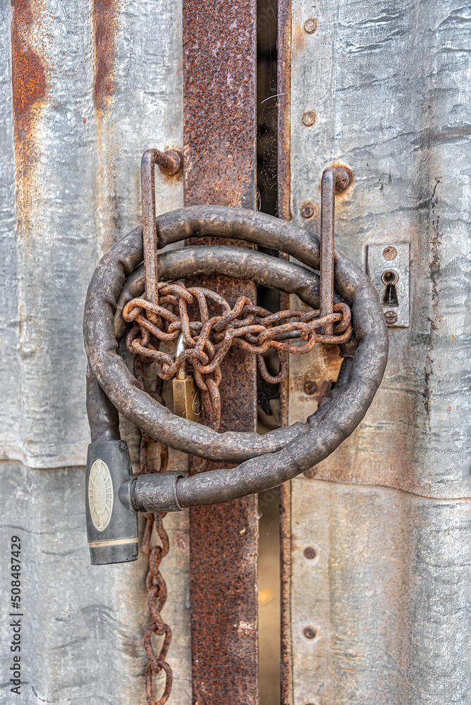 A cluster of rusty locks - old motorbike lock, rusty chain link and padlock, on rusty steel frame doors cladded with rusty corrugates metal sheets.