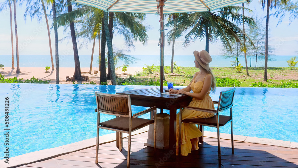 Luxury vacation in modern hotel resort. Traveler woman in dress and hat sitting in cafe near swimming pool, drinking welcome refreshment drink and enjoy tropical view with sea and palm trees