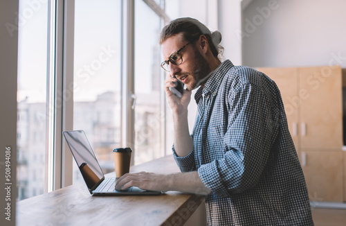 Millennial male in optical eyewear browsing website on modern laptop computer using 4g wireless for calling via mobile application, skilled programmer in spectacles using cellular gadget for talking