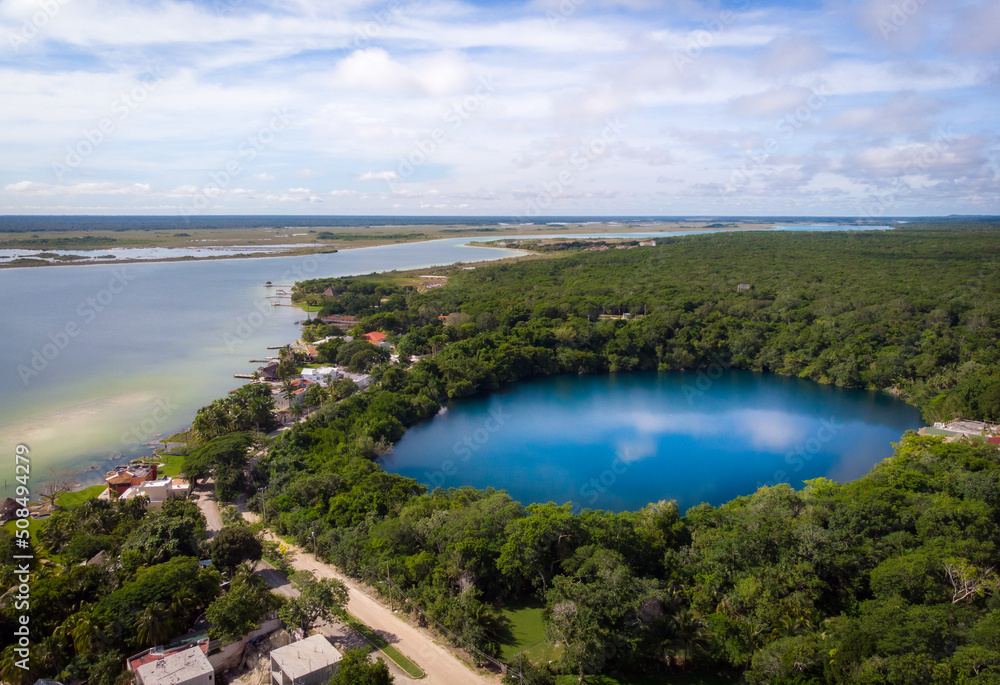 Drone view of Bacalar lagoon and cenote with blue water and light reflection surrounded by tropical green jungle on a cloudy day