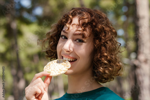 Close up view of happy redhead woman wearing green t-shirt standing on city park, outdoors looking at the camera biting healthy rice crackers smiling happy and positive.