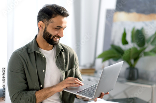 Happy attractive arabian or indian man, stock trader, real estate agent, holds laptop while stand in modern office, rejoices at the conclusion of a deal, high profits, profitable collaboration, smile
