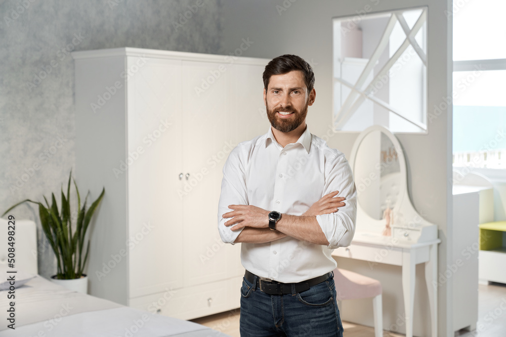 Front view of handsome seller, designer, architect standing with crossed hands in bedroom. Brunette male with beard designing, looking at camera, smiling. Concept of furniture.