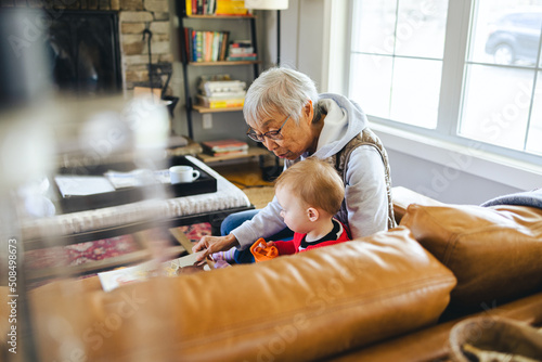 Asian grandmother reads a book to her mixed race granddaughter while sitting together on the couch