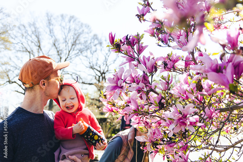 A father kisses his smiling daughter next to a magnolia tree outside in the spring time