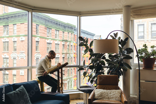 Man sits working on his digital tablet at a desk in the window with the city behind him