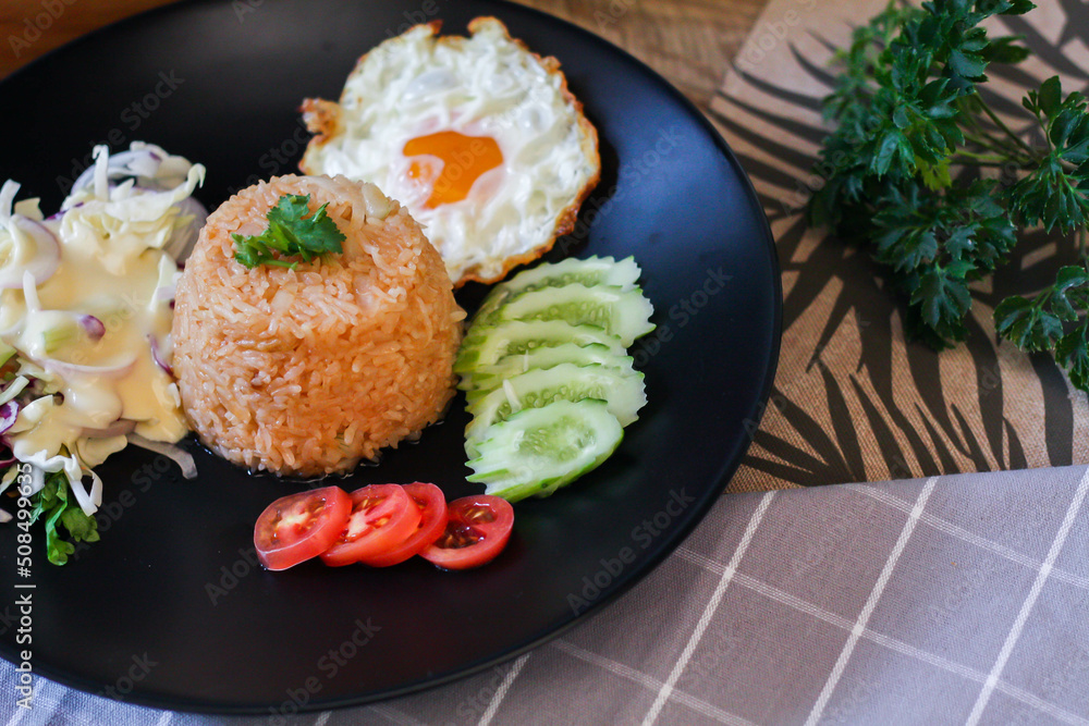 Fried rice, fried egg, cucumber, sliced tomato, green salad and Fried chicken placed on a black plate as a garnish. Fried rice, fried egg, cucumber, sliced tomato, green salad and Fried chicken placed on a black plate as a garnish.