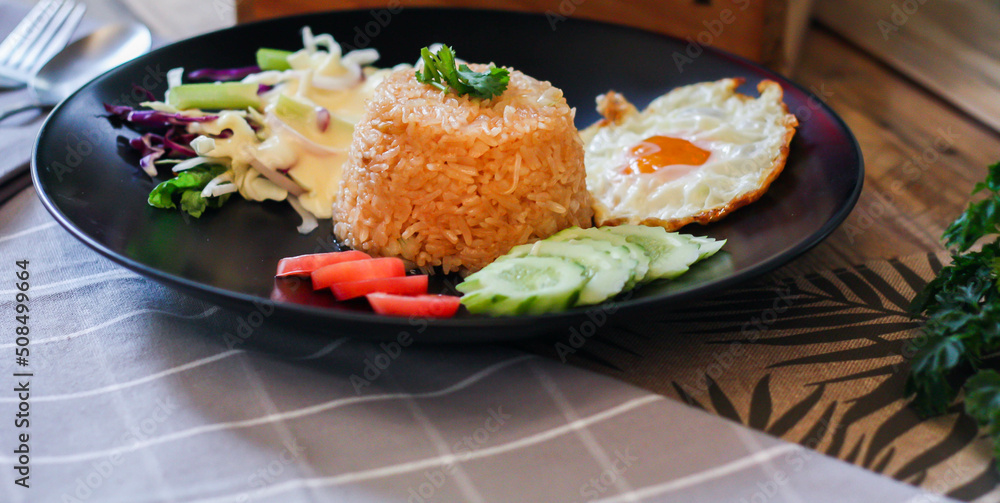 Fried rice, fried egg, cucumber, sliced tomato, green salad and Fried chicken placed on a black plate as a garnish.