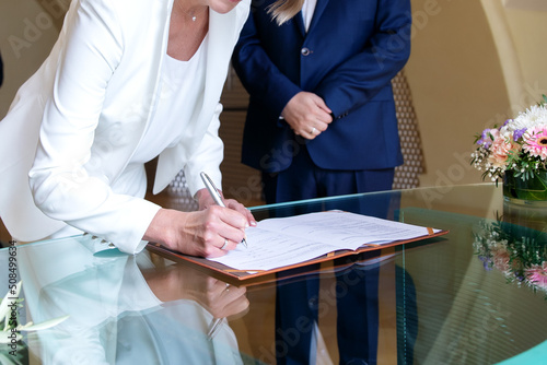 Elderly bride and groom signing marriage agreement at desk. Woman's hand holding pen and signing paper document