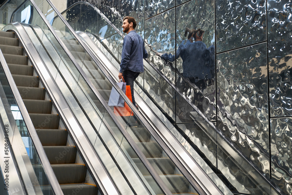 Back view of stylish young man going up by escalator. Boy with beard shopping, holding bags, purchases, smiling, looking around. Concept of modern lifestyle and shopping.