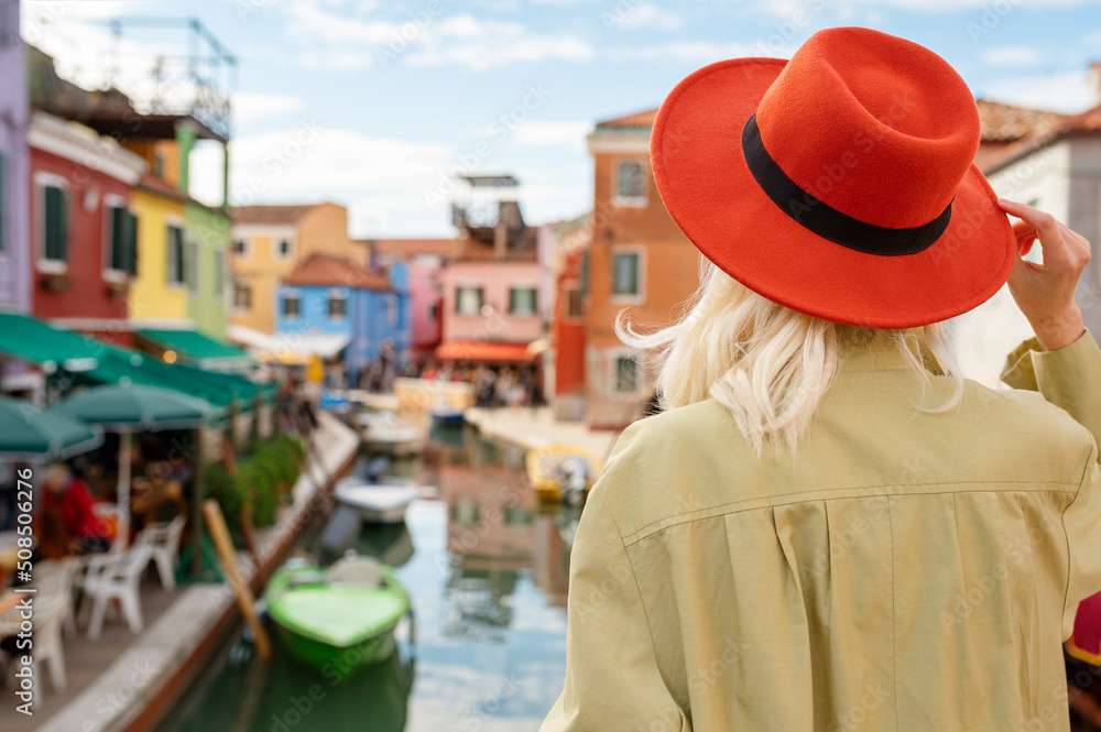 Traveler woman wearing orange hat looks at colorful houses in street of Burano island, Italy. Rear, back view. Travel, tourism, vacation, lifestyle conception. Copy, empty space for text
