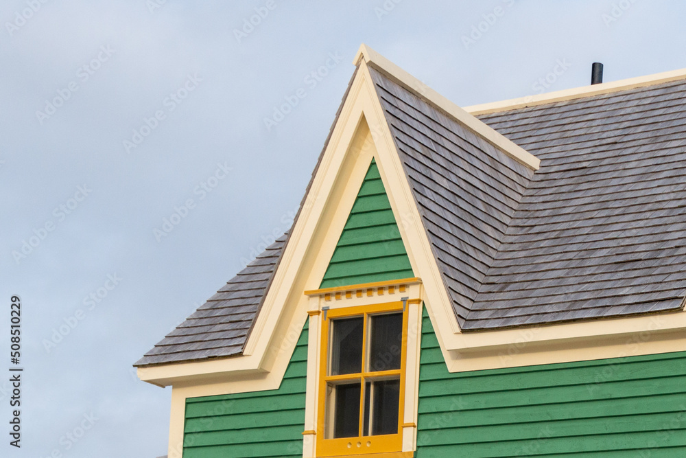 There is a small single hung window under a gable or triangle-shaped dormer on a vintage green exterior building wall with yellow trim wood case. There's a blue sky with white clouds in the background
