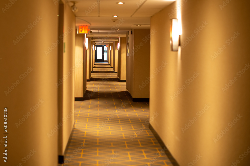  A long narrow hallway of a hotel with a red exit sign light sconce lights and cream colored walls. The carpet on the floor is green and beige with a pattern. The low ceiling has white tiles. 