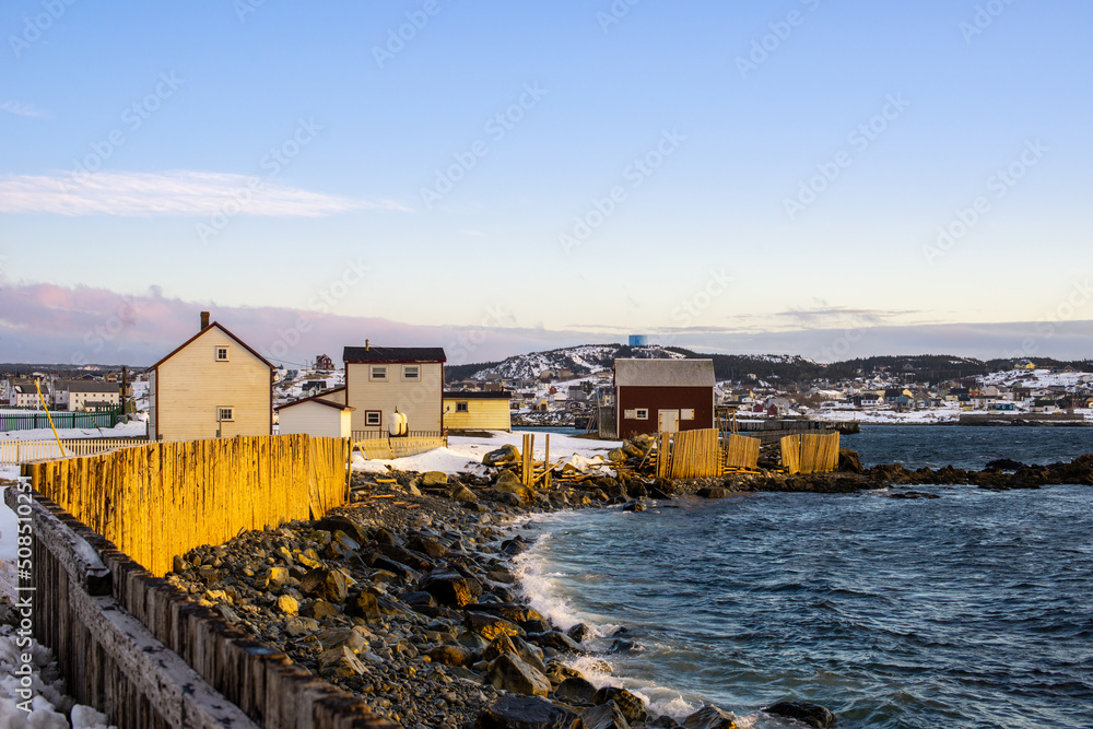 The community of Bonavista with a pale pink cloudy evening sky with a wooden seawall. Snow is piled next to the log wall. There are historic buildings at the end of the gravel road.