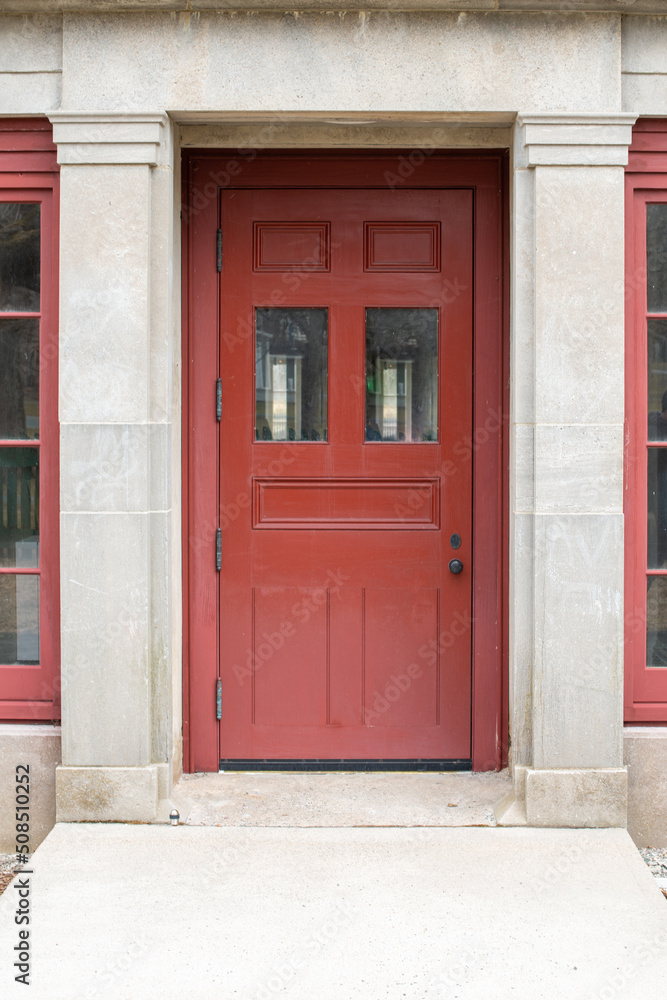 An extra wide red wooden vintage door with two small clear glass windows, multiple panels, and an old door handle. The door is in a limestone block masonry building with pillars on both sides.  