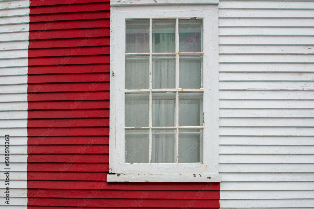 Fototapeta premium The exterior of a white wooden house wall with a red painted panel, and a closed glass single hung window. There's a white curtain in the window. The wall is made of narrow clapboard clad siding. 