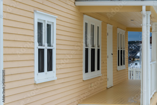 A roofed porch of a country style yellow and white house. There are multiple double hung windows and a wooden shutter door. The overhanging roof has a number of pillars and fences enclosing the deck.