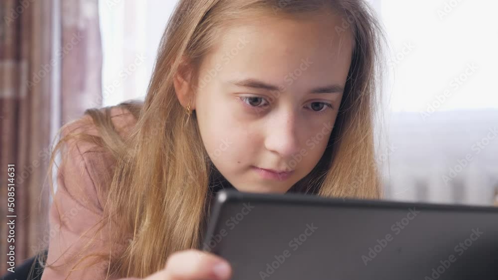 A teenage girl lies on a bed in a room with a tablet. Full face of a girl playing in a tablet, white day. A teenage girl lies on a bed in a room with a tablet. Full face of a girl playing in a tablet, white day.
