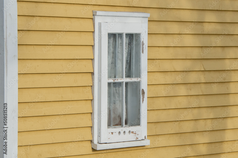 The exterior of a vintage yellow colored wall with narrow wood cape cod clapboard siding. In the center of the house, there's a closed shutter window with three air holes in the bottom. 
