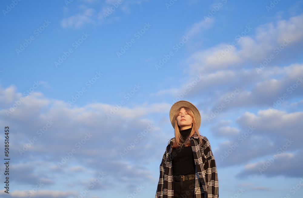 Portrait of a girl with closed eyes. Attractive blonde long hair woman in stylish clothes - shirt and brown hat on with blue cloudy sky background. Copy space for text and advertising